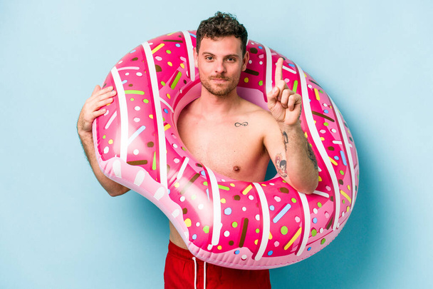 Young caucasian man holding an inflatable donut isolated on blue background showing number one with finger. - Fotografie, Obrázek