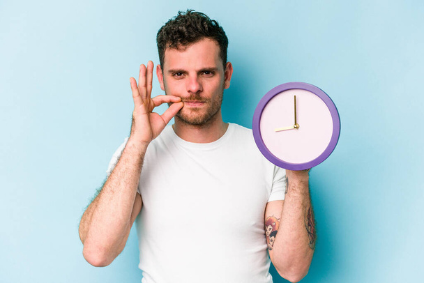 Young caucasian man holding a clock isolated on blue background with fingers on lips keeping a secret. - Foto, Bild