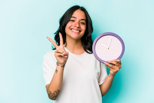Young hispanic woman holding a clock isolated on blue background showing number two with fingers. - Fotografie, Obrázek