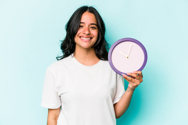 Joven mujer hispana sosteniendo un reloj aislado sobre fondo azul feliz, sonriente y alegre. - Foto, imagen