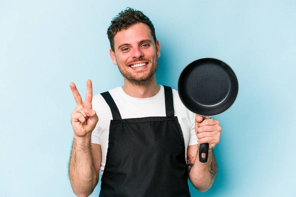 Jeune cuisinière caucasienne tenant poêle isolée sur fond bleu montrant numéro deux avec les doigts. - Photo, image