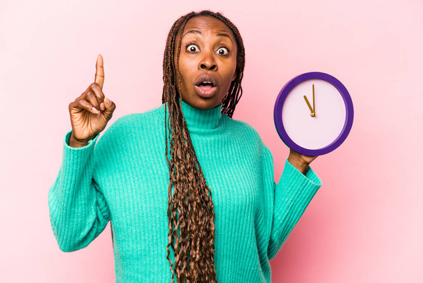 Young African American woman holding a clock isolated on pink background having an idea, inspiration concept. - Photo, Image