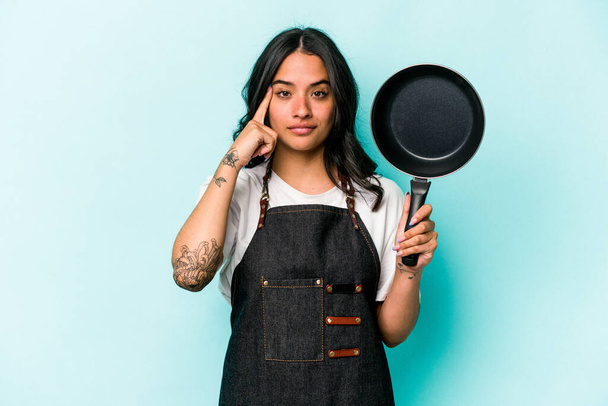 Jeune cuisinière hispanique femme tenant poêle isolée sur fond bleu pointant temple avec le doigt, penser, concentré sur une tâche. - Photo, image