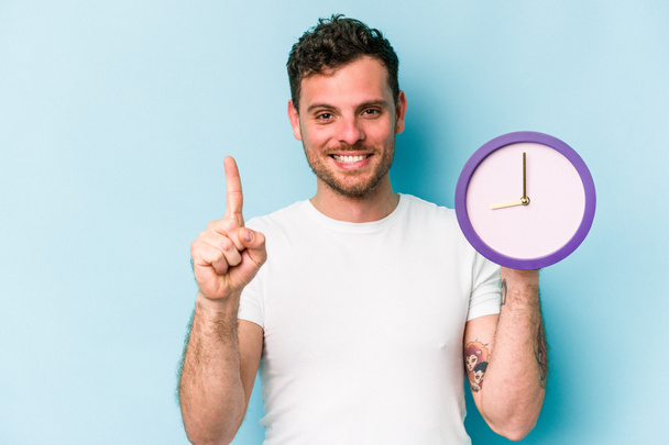 Young caucasian man holding a clock isolated on blue background showing number one with finger. - Photo, image