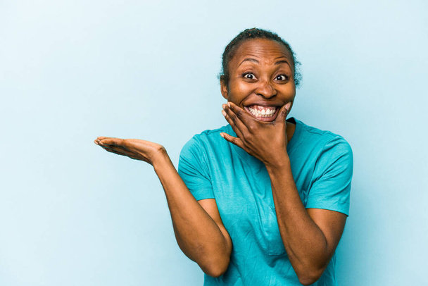 Νεαρή Αφρικάνα Αμερικανίδα, απομονωμένη σε μπλε φόντο έχει χώρο για φωτοτυπίες σε μια παλάμη, κρατώντας το χέρι πάνω από το μάγουλο. Έκπληκτη και χαρούμενη. - Φωτογραφία, εικόνα