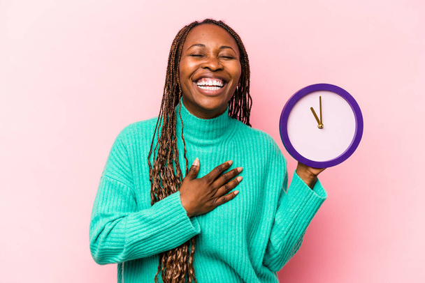 Joven mujer afroamericana sosteniendo un reloj aislado sobre fondo rosa se ríe en voz alta manteniendo la mano en el pecho. - Foto, imagen