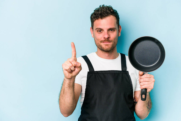 Young caucasian cooker holding frying pan isolated on blue background showing number one with finger. - Foto, immagini