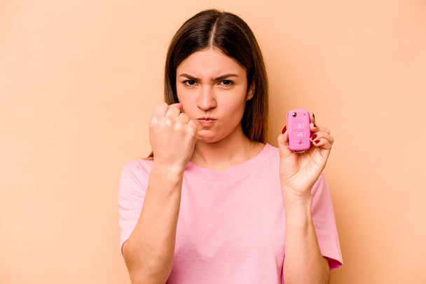 Giovane donna ispanica in possesso di una chiave auto isolata su sfondo beige mostrando pugno alla macchina fotografica, espressione facciale aggressiva. - Foto, immagini