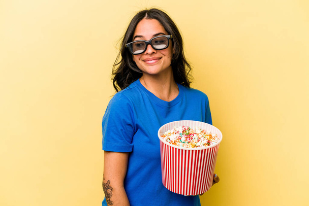 Young hispanic woman holding popcorn isolated on yellow background looks aside smiling, cheerful and pleasant. - Фото, изображение