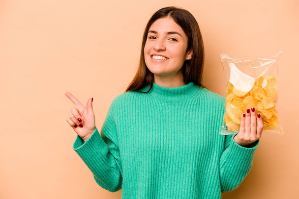Mujer hispana joven sosteniendo una bolsa de papas fritas aisladas sobre fondo beige sonriendo y señalando a un lado, mostrando algo en espacio en blanco. - Foto, imagen