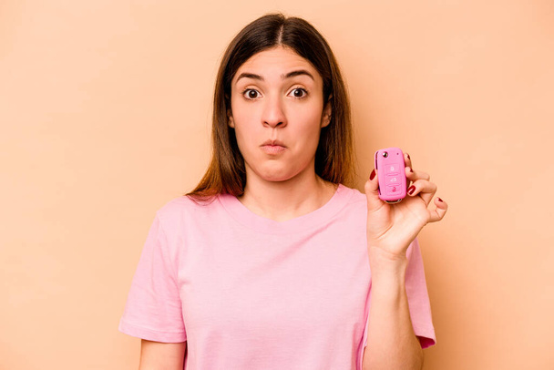 Young hispanic woman holding a keys car isolated on beige background shrugs shoulders and open eyes confused. - Photo, Image