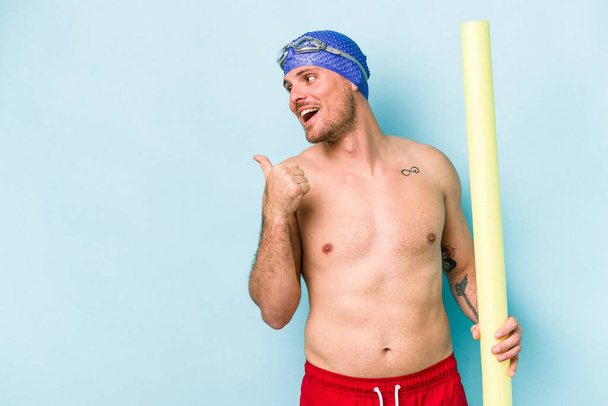 Young caucasian swimmer man holding foam stick isolated on blue background points with thumb finger away, laughing and carefree. - Fotografie, Obrázek