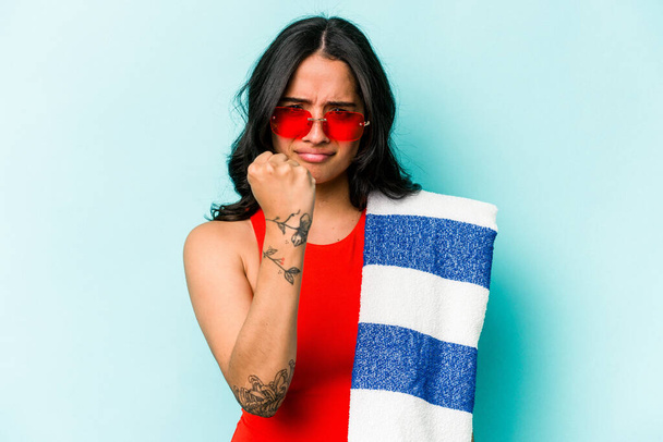 Young hispanic woman holding beach towel isolated on blue background showing fist to camera, aggressive facial expression. - 写真・画像