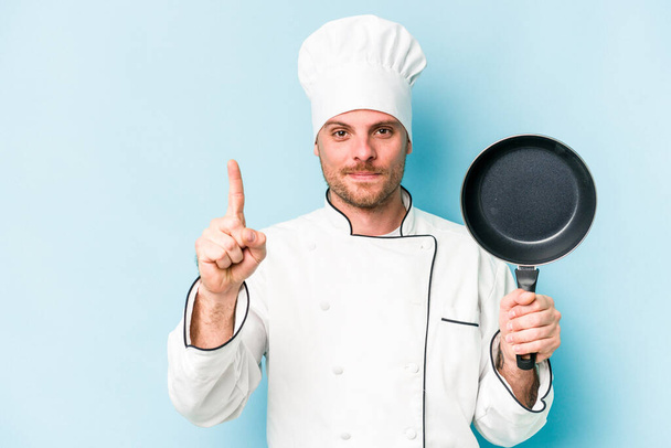Young caucasian chef man holding flying pan isolated on blue background showing number one with finger. - Valokuva, kuva