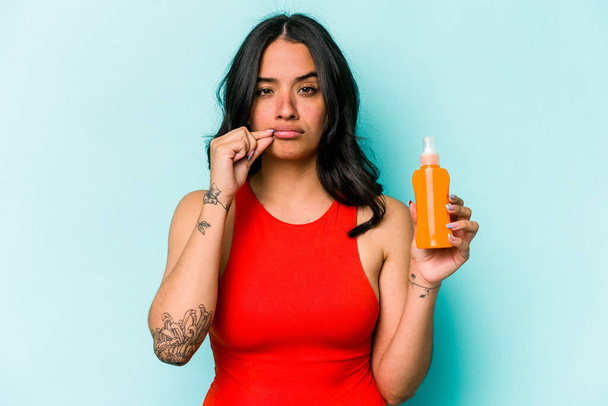 Young hispanic woman holding sun cream isolated on blue background with fingers on lips keeping a secret. - Photo, Image