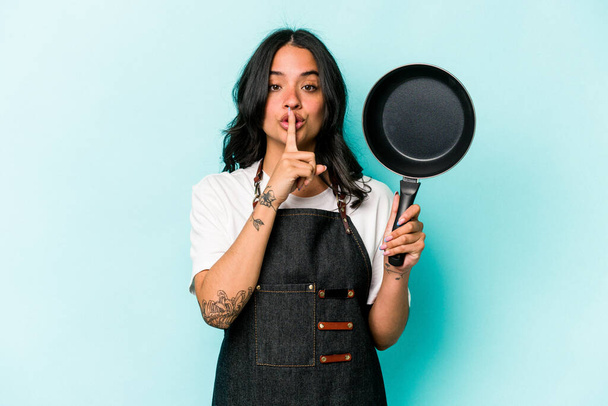 Young hispanic cooker woman holding frying pan isolated on blue background keeping a secret or asking for silence. - 写真・画像