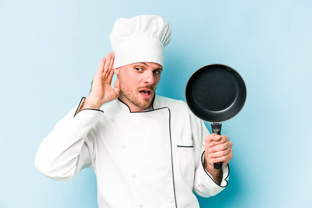 Young caucasian chef man holding flying pan isolated on blue background trying to listening a gossip. - Φωτογραφία, εικόνα