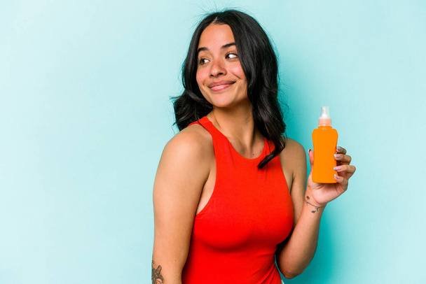 Young hispanic woman holding sun cream isolated on blue background looks aside smiling, cheerful and pleasant. - Foto, Bild