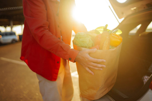 Healthy vegan vegetarian food in a paper bag  in the male hands. Young man with shopping bag near the car. Consumerism, sale, purchases, shopping, lifestyle concept. - Foto, imagen