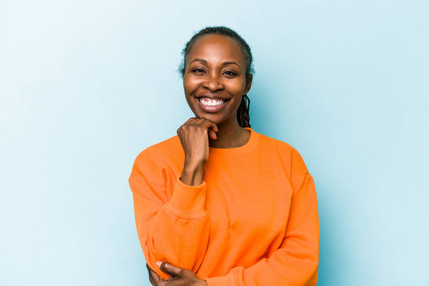 Νεαρή Αφροαμερικανή γυναίκα απομονωμένη σε μπλε φόντο χαμογελώντας χαρούμενη και σίγουρη, αγγίζοντας το πηγούνι με το χέρι. - Φωτογραφία, εικόνα