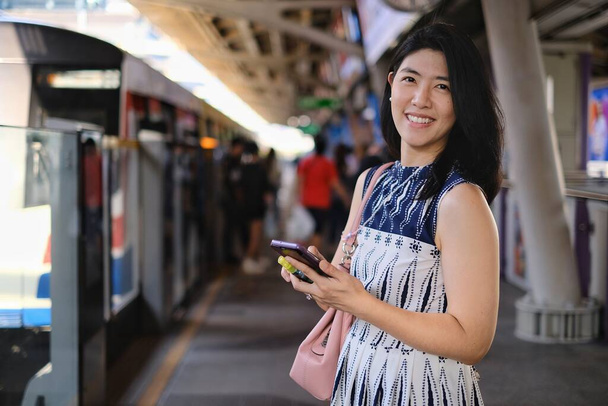 Une jolie jeune femme d'affaires asiatique confiante utilise son téléphone portable en attendant son train dans une gare pour aller travailler le matin. - Photo, image