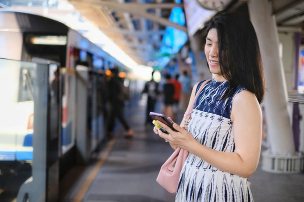 Μια ελκυστική νεαρή σίγουρη Ασιάτισσα επιχειρηματίας χρησιμοποιεί το κινητό της ενώ περιμένει το τρένο της σε ένα σταθμό να πάει στη δουλειά το πρωί.. - Φωτογραφία, εικόνα