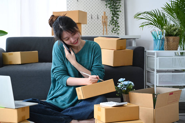 Mujer asiática sonriente vendedor en línea recibe orden en el teléfono móvil y la preparación de cajas de paquete de producto para la entrega - Foto, Imagen
