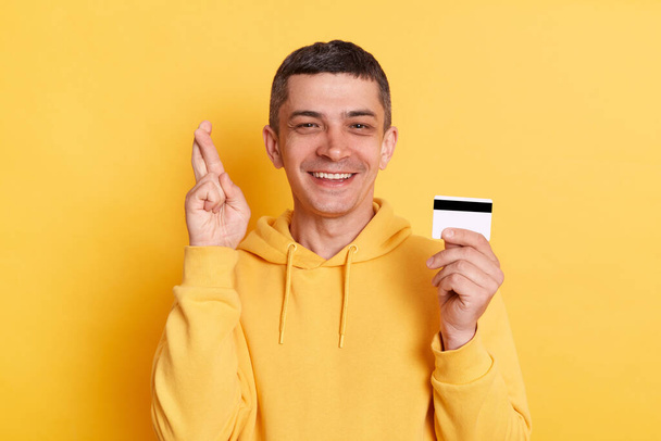 Portrait of delighted optimistic man wearing casual style hoodie, posing isolated over yellow background, holding credit card and crossing finger, hopes to get money on account. - Fotó, kép