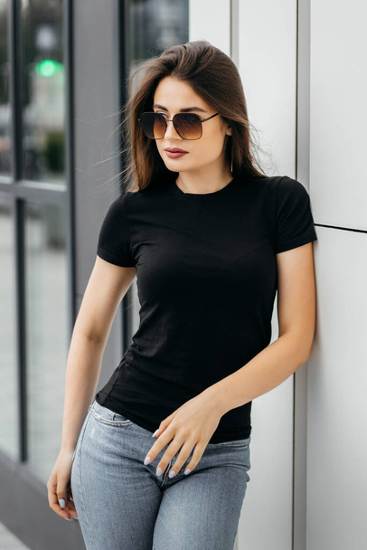 Elegante ragazza bruna che indossa t-shirt nera e occhiali da sole in posa contro strada, stile di abbigliamento urbano. Foto di strada - Foto, immagini