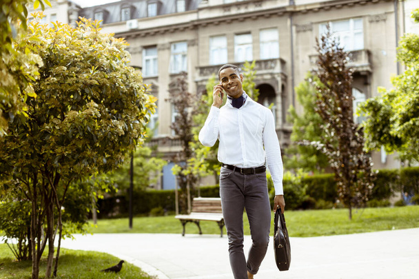 Knappe jonge Afro-Amerikaanse zakenman met behulp van een mobiele telefoon tijdens het lopen op straat - Foto, afbeelding