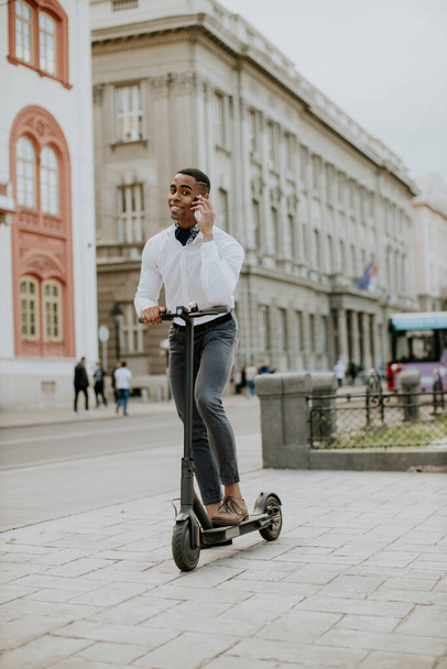 Sokakta elektrikli scooterla dururken cep telefonu kullanan yakışıklı Afrikalı genç bir Amerikalı. - Fotoğraf, Görsel