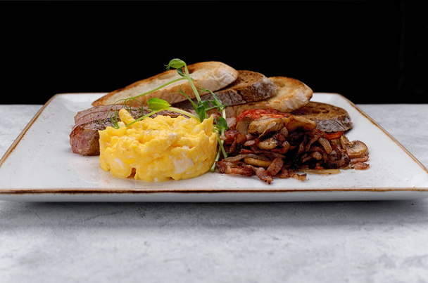 Irish breakfast on a plate. Sausage, scrambled eggs, mushrooms, bacon, ciabatta toast. On a mixed background - Photo, Image
