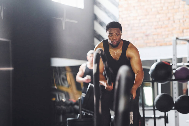 Strong african american man in sportive clothes have workout day in the gym. Doing crossfit by using ropes. - Foto, Imagem