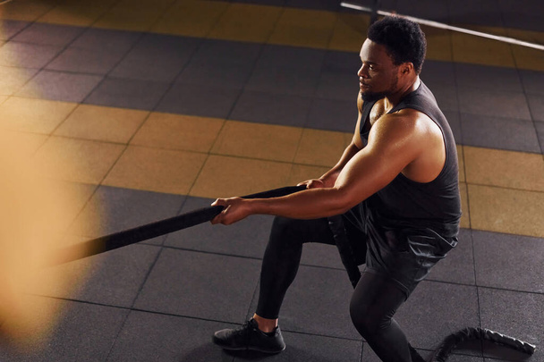 Strong african american man in sportive clothes have workout day in the gym. Doing crossfit by using ropes. - 写真・画像