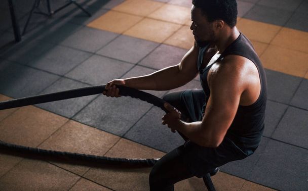 Strong african american man in sportive clothes have workout day in the gym. Doing crossfit by using ropes. - Φωτογραφία, εικόνα
