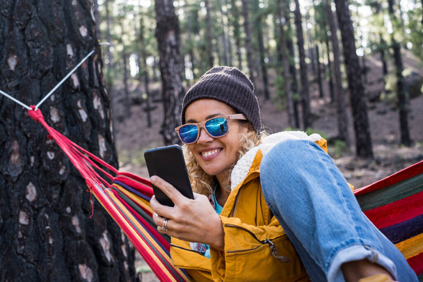 Happy woman in video call conference with phone on hammock in the woods forest outdoor park - vidám emberek és szabadság alternatív életmód és utazás - Fotó, kép