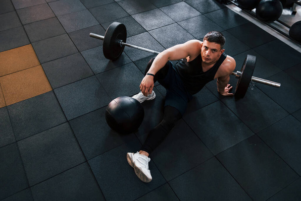 Strong man in sportive clothes sitting on the floor in the gym with equipment. - 写真・画像
