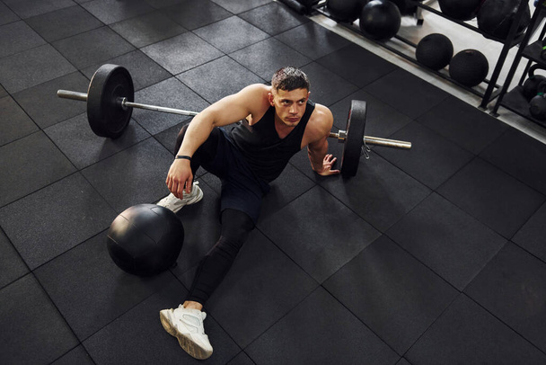 Strong man in sportive clothes sitting on the floor in the gym with equipment. - Foto, afbeelding