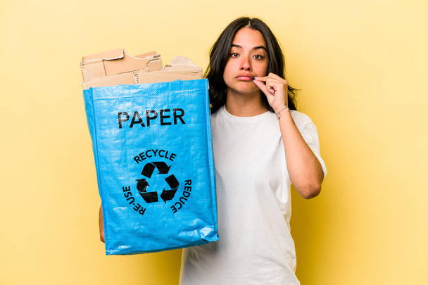 Молодая латиноамериканка перерабатывает бумагу на желтом фоне с пальцами на губах, хранящими секрет. - Фото, изображение