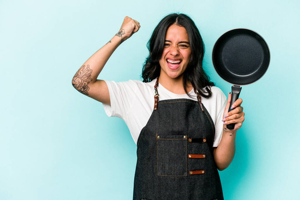 Joven cocinera hispana sosteniendo sartén aislada sobre fondo azul levantando puño después de una victoria, concepto ganador. - Foto, imagen
