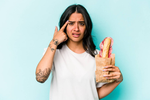 Młoda Latynoska kobieta jedząca kanapkę odizolowaną na niebieskim tle wykazującą rozczarowanie palcem wskazującym. - Zdjęcie, obraz