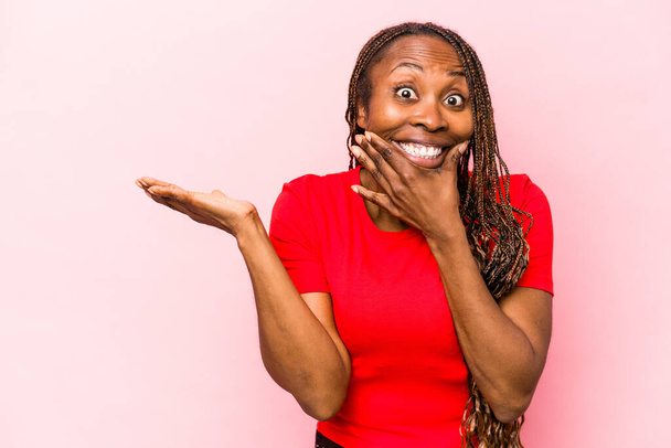 Junge afrikanisch-amerikanische Frau isoliert auf rosa Hintergrund hält Kopierraum auf einer Handfläche, halten Hand über Wange. Erstaunt und erfreut. - Foto, Bild