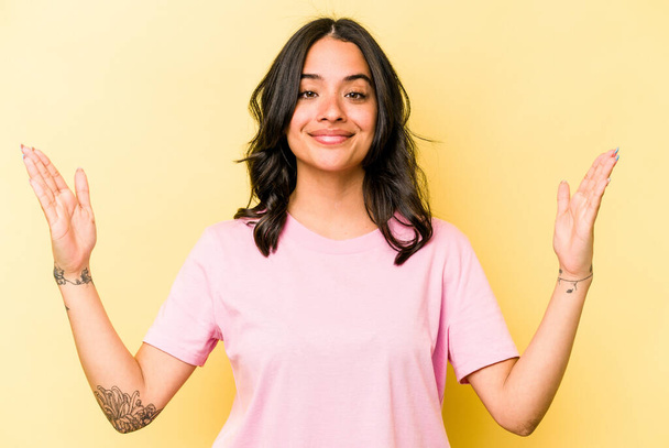 jonge Spaanse vrouw geïsoleerd op gele achtergrond houden iets weinig met wijsvingers, glimlachend en vol vertrouwen. - Foto, afbeelding