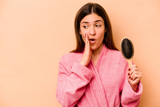 Young hispanic woman holding hairbrush isolated on beige background is saying a secret hot braking news and looking aside - Fotó, kép