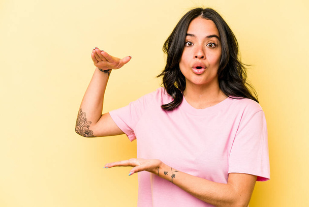 Young hispanic woman isolated on yellow background shocked and amazed holding a copy space between hands. - Photo, image