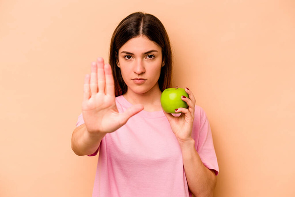 Young hispanic woman holding an apple isolated on beige background standing with outstretched hand showing stop sign, preventing you. - Fotoğraf, Görsel