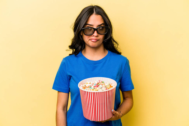 Young hispanic woman holding popcorn isolated on yellow background confused, feels doubtful and unsure. - 写真・画像
