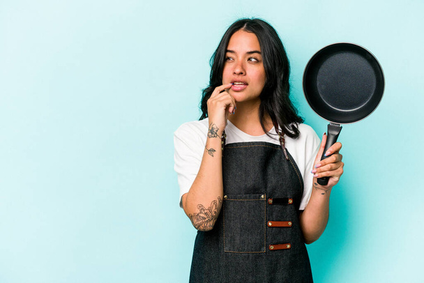 Молодая латиноамериканка держит сковородку, изолированную на синем фоне, расслабленно думая о чем-то, глядя на пространство для копирования.. - Фото, изображение