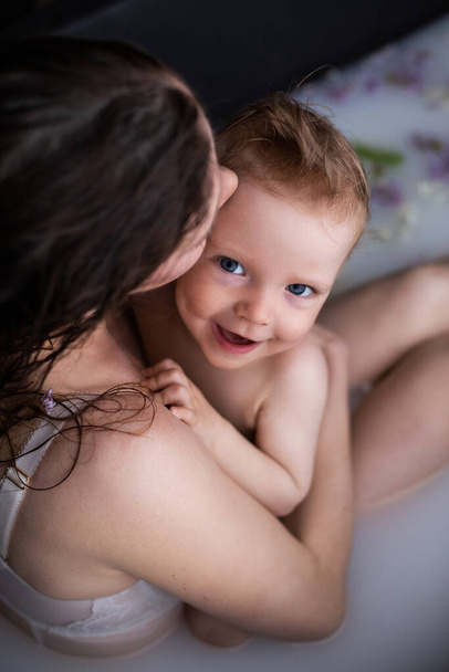Νεαρή ευτυχισμένη μητέρα με τον μικρό γιο της σε μπάνιο με ανθόγαλα. Η μαμά και το παιδί αγκαλιάζονται στο μπάνιο. - Φωτογραφία, εικόνα