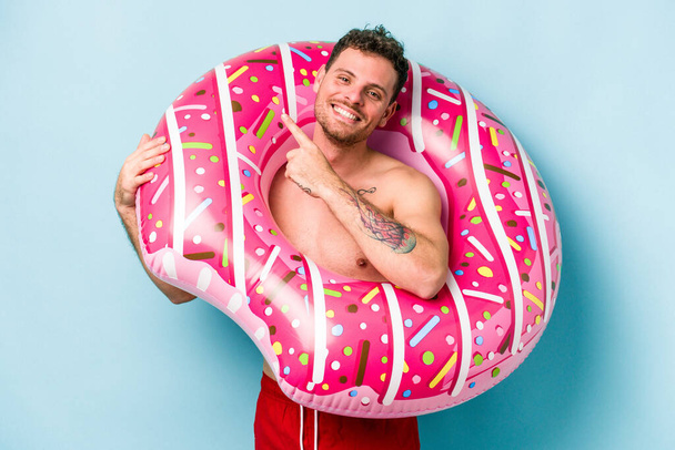 Young caucasian man holding an inflatable donut isolated on blue background smiling and pointing aside, showing something at blank space. - Foto, Bild